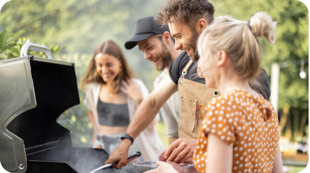 A man cooking meat on a barbeque while his friends look on happily. 