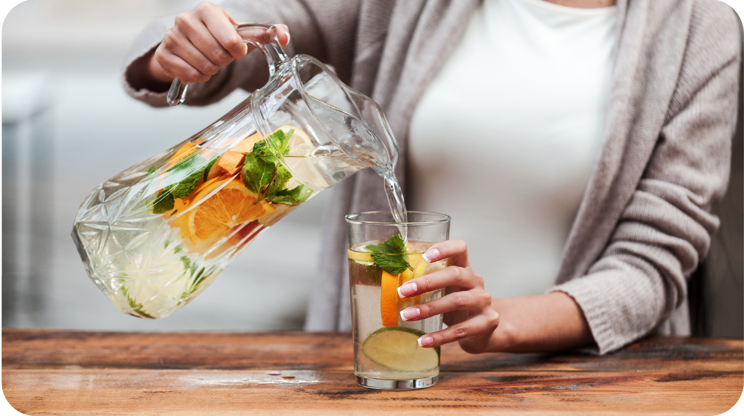 A close-up of a woman's hands pouring a jug filled with water and fruits into a glass. 