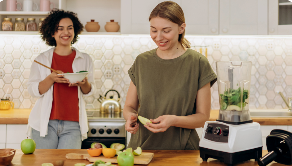 A woman cutting fruits and vegetables smiling with another woman in the background looking happy