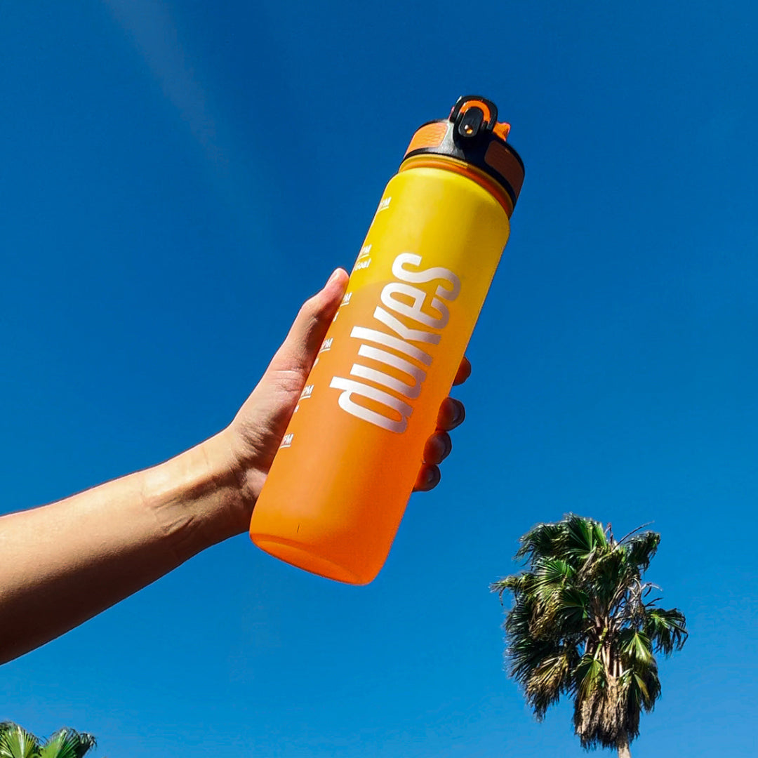 A close-up Dukes motivational bottle being held up with a blue sky background and a palm tree on the bottom right.