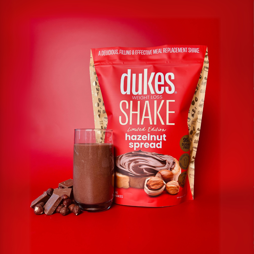 A bag of Dukes Weight Loss Shake Hazelnut Spread next to a glass of the shake, chocolate and hazelnuts